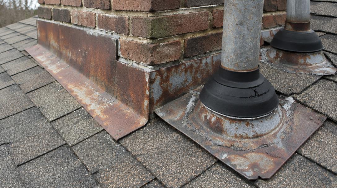 Rusted step flashing and cracked pipe boots beside a brick chimney on an asphalt shingle roof